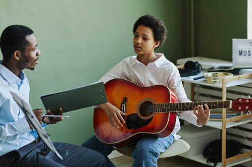 A black boy and a guitar teacher talking during a guitar lesson, with the boy holding an acoustic guitar.