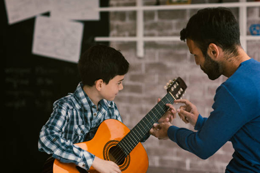 A guitar teacher is explaining something to a boy, pointing to the guitar neck, while the boy holds the guitar.